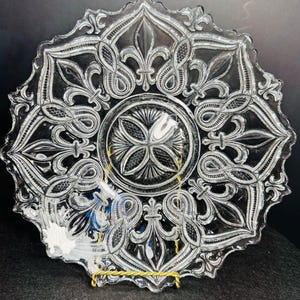 May include: A clear pressed glass plate featuring a central circular medallion with a cross-hatched pattern, surrounded by a repeating border of fleur-de-lis motifs and intricate scrollwork, finished with a scalloped, decorative edge.