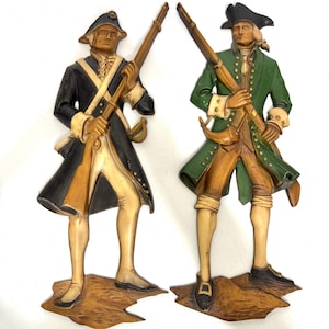 May include: Two wooden wall hangings depicting colonial soldiers. One soldier wears a black coat with gold accents, the other a green coat. Both figures hold rifles and stand on a brown base. The figures are painted in shades of brown, beige, black, and green.