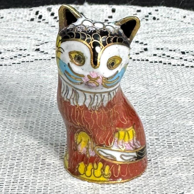 May include: A decorative cat figurine with a red body, white face, and gold accents. The cat's face features yellow eyes, blue accents, and a pink nose. The figurine has a detailed design and is likely made of ceramic or porcelain.