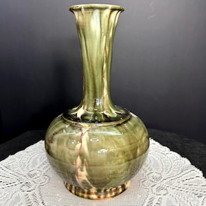 May include: A green ceramic vase with a bulbous base and a tall, fluted neck. The vase has a glossy finish with streaks of brown and cream. The vase is a decorative item, suitable for displaying flowers or as a standalone art piece.