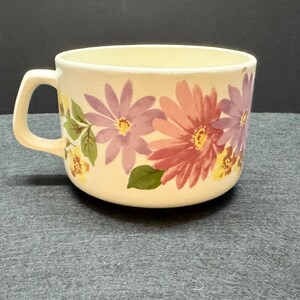 May include: Cream-colored ceramic mug with a floral design. The mug features a handle and is decorated with large pink, purple, and yellow flowers with green leaves. The mug is likely used for drinking beverages.