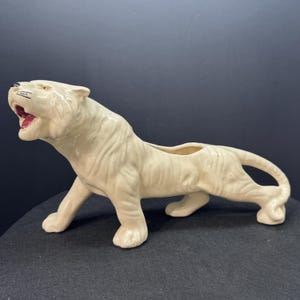 May include: Cream-colored ceramic tiger figurine with an open mouth, revealing a red interior. The tiger is in a walking pose, with a planter opening on its back. The figurine has detailed fur texture and is suitable for home decor.