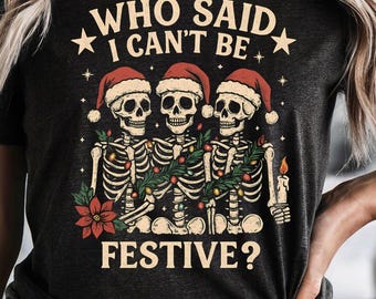 May include: Dark gray t-shirt featuring a festive skeleton design. The graphic includes three skeletons wearing Santa hats and holding garland, with the text "WHO SAID I CAN'T BE FESTIVE?" in a vintage style. A candle and poinsettia are also included.