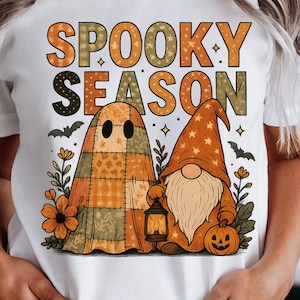 Könnte beinhalten: Weißes T-Shirt mit den Worten "Spooky Season" in orangefarbenen und grünen Blockbuchstaben. Das Shirt zeigt einen Cartoon-Geist und einen Zwerg mit einem Kürbis und einer Laterne, perfekt für Halloween.