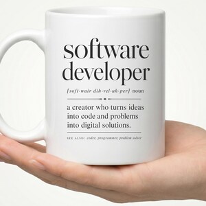 May include: A white ceramic mug featuring black printed text that defines a software developer as a creator who turns ideas into code and problems into digital solutions, including phonetic pronunciation and related terms like coder and programmer.