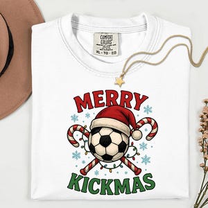 May include: White t-shirt with a Christmas-themed soccer ball graphic. The design features a soccer ball wearing a Santa hat, candy canes, and the words "MERRY KICKMAS" in red and green. A gold necklace with a star pendant rests on the shirt.