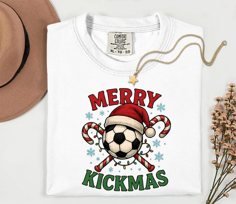 May include: White t-shirt with a Christmas-themed soccer ball graphic. The design features a soccer ball wearing a Santa hat, candy canes, and the words "MERRY KICKMAS" in red and green. A gold necklace with a star pendant rests on the shirt.