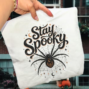 White t-shirt with "Stay Spooky" text, spider on a web, and Halloween decor.