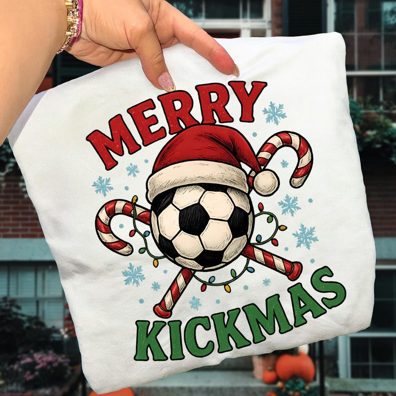 May include: White pillow with a festive design. The design features a soccer ball wearing a Santa hat, candy canes, snowflakes, and the words "MERRY KICKMAS" in red and green. A string of lights is wrapped around the candy canes.