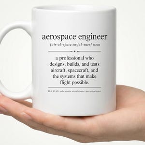 May include: A white ceramic mug featuring black printed text that defines the term aerospace engineer as a professional who designs, builds, and tests aircraft, spacecraft, and the systems that make flight possible, including phonetic pronunciation.