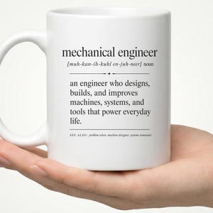 May include: A white ceramic mug featuring black printed text that defines a mechanical engineer as an engineer who designs, builds, and improves machines, systems, and tools that power everyday life, including phonetic pronunciation and see also.