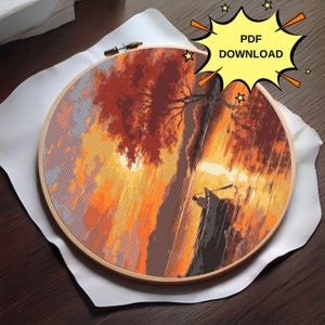 K&ouml;nnte beinhalten: Ein runder Holzstickrahmen mit einem farbenfrohen Kreuzstich-Design, das eine Sonnenuntergangsszene mit einem Boot und einem Baum darstellt. Das Design ist in Orange-, Gelb-, Rot- und Braunt&ouml;nen gehalten.