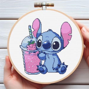 May include: Cross-stitch embroidery featuring a blue cartoon character with large eyes and pink ears, holding a pink strawberry smoothie. The artwork is set in a wooden hoop, suitable for home decor or a handmade gift.