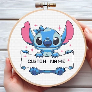 Op de afbeelding: Een borduurring met kruissteek met een blauw stripfiguur met grote oren en een strik, met een banner met de tekst "CUSTOM NAME". Het personage is omgeven door roze steraccenten, tegen een witte achtergrond.