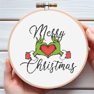 May include: A cross-stitch embroidery hoop with the words "Merry Christmas" in black script. The design features green hands forming a heart around a red heart. The hoop is made of wood and is held by hands.