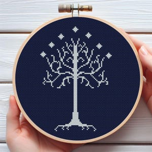 May include: A white cross-stitch of a tree with star-shaped accents on a navy blue background, set in a wooden embroidery hoop. The tree design is centered, with a detailed trunk and branches.