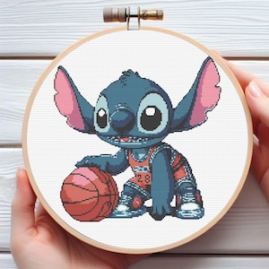 Puede incluir: Arte de aro de punto de cruz con el personaje Stitch de Lilo & Stitch, botando un balón de baloncesto. Stitch lleva una camiseta de baloncesto con el número 28. La obra está enmarcada en un aro de bordado de madera.
