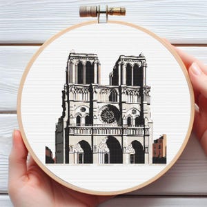 May include: A black and white cross-stitch embroidery of Notre Dame Cathedral in Paris, France. The embroidery is framed in a round wooden hoop.