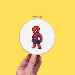 May include: A cross-stitch embroidery hoop featuring a pixelated Spider-Man design. The superhero is depicted in his classic red and blue suit, with a white background and a wooden hoop. The artwork is held against a yellow background.