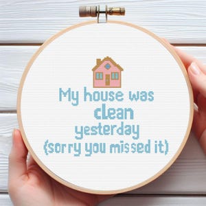 Könnte beinhalten: Ein Kreuzstich-Stickrahmen mit dem Text "My house was clean yesterday (sorry you missed it)". Das Design zeigt ein rosa Haus und blauen Text auf weißem Hintergrund, in einem Holzrahmen.