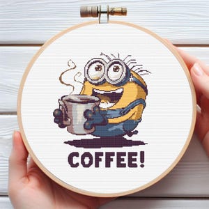 Może przedstawiać: Haft krzyżykowy w drewnianym tamborku przedstawiający żółtą postać Minionka trzymającą filiżankę kawy. Słowo "COFFEE!" jest wyhaftowane poniżej. Wzór znajduje się na białej tkaninie.