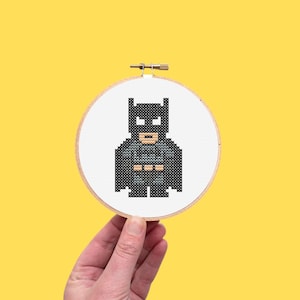 May include: A cross-stitch embroidery hoop featuring a pixelated Batman design. The superhero is depicted in shades of black, gray, and white, set against a white fabric background. The hoop is held against a yellow background.