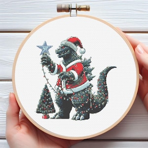 Puede incluir: Arte de aro de punto de cruz que presenta a Godzilla con un traje de Papá Noel, sosteniendo una estrella y decorando un árbol de Navidad. El diseño está sobre tela blanca, dentro de un aro de madera. La obra es festiva y navideña.