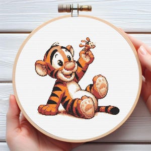 Puede incluir: Bordado de punto de cruz de un tigre de dibujos animados sosteniendo un pequeño juguete. El tigre es naranja y negro, con una barriga blanca y una expresión amigable. El bordado está en un aro de madera.