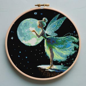 May include: A cross-stitch embroidery of a fairy holding a glowing orb in front of a large moon. The fairy is wearing a green dress and has translucent wings. The background is black with small white stars, all within a wooden embroidery hoop.