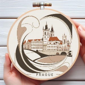 May include: A round embroidery hoop with a beige background and a brown and beige illustration of the Prague skyline. The illustration features a bridge, buildings, and a decorative swirl design. The text "PRAGUE" is printed in brown at the bottom of the hoop.