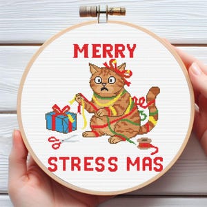 May include: A round wooden embroidery hoop containing a cross-stitch design of a brown tabby cat tangled in red, yellow, and green ribbons. The text MERRY STRESS MAS is stitched in red. A blue gift box and red scissors are also depicted.