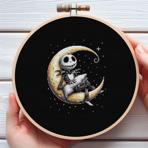 May include: A cross-stitch embroidery hoop with a black background and a design of a skeleton character on a crescent moon. The moon is yellow and the character is black and white. The hoop is held by hands.