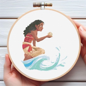 Op de afbeelding: Kruissteekborduurwerk in een houten hoepel. Het ontwerp toont een Polynesische vrouw die een zeeschelp vasthoudt, met oceaangolven op de voorgrond. De vrouw draagt een rode en beige outfit. De achtergrond is wit.