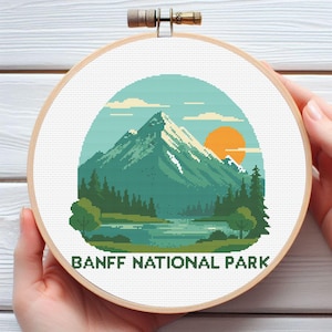 May include: A cross-stitch embroidery hoop featuring a landscape design of Banff National Park. The design includes a mountain, lake, trees, and a setting sun, all in shades of green, blue, and orange. The text "BANFF NATIONAL PARK" is at the bottom.