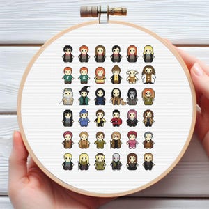 Puede incluir: Bordado de punto de cruz con personajes pixelados en un aro de madera. Los personajes están representados en un estilo de dibujos animados, con una variedad de colores y atuendos. El bordado está sobre un fondo de tela blanca.