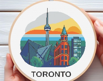 May include: A cross-stitch embroidery hoop featuring a colorful Toronto cityscape. The design includes the CN Tower, a red brick building, and the word "TORONTO" in black. The background has a sunset gradient of orange, yellow, and blue.
