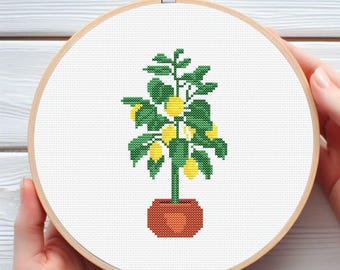 May include: A cross-stitch embroidery of a lemon tree in a terracotta pot, set in a wooden hoop. The tree features green leaves and yellow lemons. The embroidery is held by a person's hands.