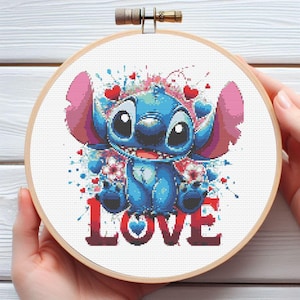 Può includere: Un telaio da ricamo a punto croce con un personaggio blu dei cartoni animati con grandi occhi e orecchie rosa, circondato da cuori e fiori. La parola "LOVE" è ricamata in rosso in basso. Il telaio è tenuto da una cornice di legno.