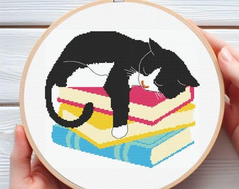 May include: A cross-stitch embroidery hoop featuring a black and white cat sleeping on a stack of colorful books. The books are in shades of pink, yellow, and blue. The cat has a white chest and paws, and a pink nose.