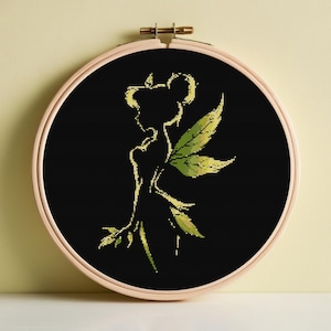 Puede incluir: Bordado de punto de cruz de una silueta de hada con alas y acentos de hojas verdes, sobre un fondo negro. El diseño está enmarcado en un bastidor de bordado redondo de madera.