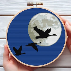 May include: A cross-stitch embroidery hoop with a blue background and a large white moon. Three black silhouettes of birds are flying in front of the moon.