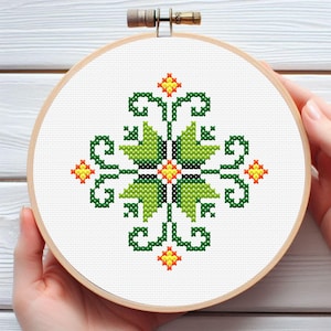 Könnte beinhalten: Fertige Kreuzstich-Stickerei in einem Holzrahmen. Das Design zeigt ein symmetrisches Blumenmuster in Grün-, Gelb- und Orangetönen auf weißem Hintergrund. Die Stickerei wird von den Händen einer Person gehalten.