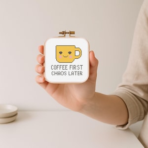 May include: A small, square cross-stitch embroidery hoop featuring a yellow coffee cup with a smiling face. The text "COFFEE FIRST CHAOS LATER" is stitched below the cup in black letters. The hoop is held in a hand.