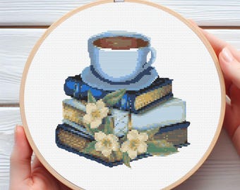 May include: A white coffee cup with a brown liquid inside sits on a stack of books with a blue cover. The books are surrounded by white flowers.
