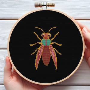 May include: A cross-stitch embroidery hoop with a black background and a colourful insect design. The insect has a red body, yellow wings, and a teal head.