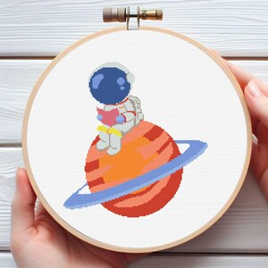 Puede incluir: Bordado de punto de cruz en un aro de madera que representa a un astronauta de dibujos animados leyendo un libro en un planeta con anillos. El astronauta lleva un casco azul y un traje espacial blanco. El planeta es naranja y rojo con un anillo azul.