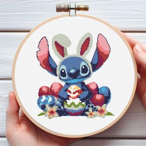 May include: A cross-stitch embroidery hoop featuring a blue cartoon character wearing a bunny ear headband and holding a decorated Easter egg. The design includes red and blue Easter eggs and flowers, all set against a white background.