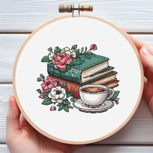 May include: Cross-stitch embroidery in a wooden hoop featuring a stack of books, pink and white roses, and a teacup with saucer. The books are green and brown, and the teacup is white with a brown beverage.