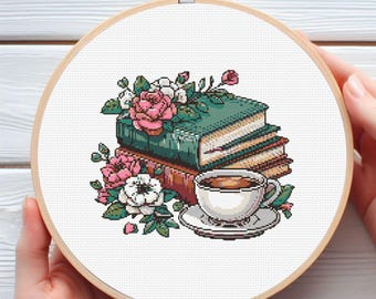 May include: Cross-stitch embroidery in a wooden hoop featuring a stack of books, pink and white roses, and a teacup with saucer. The books are green and brown, and the teacup is white with a brown beverage.