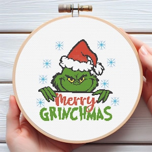Pode incluir: Um bastidor de bordado em ponto cruz com o Grinch usando um gorro de Papai Noel. O Grinch é verde com olhos amarelos e o texto "Merry Grinchmas" em vermelho e verde. Flocos de neve azuis estão espalhados.